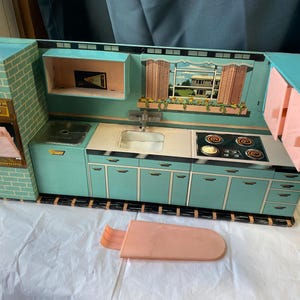 May include: A vintage toy kitchen set in teal, pink, and white. The set features a stove, sink, cabinets, and a window with a scenic view. The oven door is open, and a pink accessory is in the foreground.