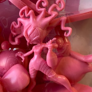 May include: A monochromatic pink plastic figurine featuring a stylized character with long, wavy, tentacle-like hair and a slender build. The figure is posed dynamically with one arm raised, surrounded by other pink molded plastic toy components.
