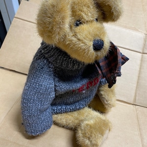May include: A plush dog toy with tan fur and a gray knit sweater. The sweater has red lettering and the dog has a plaid scarf. The dog is sitting on a brown cardboard surface.