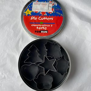 Peut inclure: Un ensemble d'emporte-pièces à tarte dans une boîte ronde. Le couvercle de la boîte est rouge et bleu avec les mots "Pie Cutters" et "emporte-pièces à tarte". À l'intérieur, plusieurs emporte-pièces en métal noir de différentes formes.