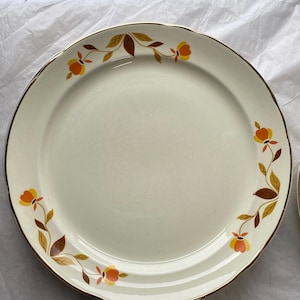 Può includere: Un piatto piano rotondo in ceramica color bianco sporco con bordo smerlato e un sottile profilo dorato. Il piatto è decorato con un motivo ripetuto di fiori arancioni e gialli accompagnati da foglie marroni e beige lungo il bordo esterno.