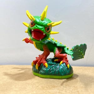 May include: A green and yellow dragon figurine with a red mouth and claws. The dragon has yellow spikes and is posed on a green base. The figurine is a toy or collectible item.