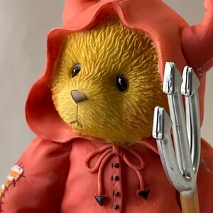 May include: A yellow teddy bear figurine wearing a red devil costume with a hood and horns. The bear holds a silver pitchfork. The costume has a bow at the neck and is buttoned down the front. The bear has black eyes and a brown nose.