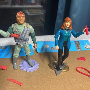 Puede incluir: Dos figuras de acción de Star Trek. Una figura es un alienígena masculino con uniforme verde y gris que sostiene un arma roja. La otra figura es una mujer con una chaqueta azul y pantalones negros, también sosteniendo un arma roja. Varios accesorios rojos están dispersos.