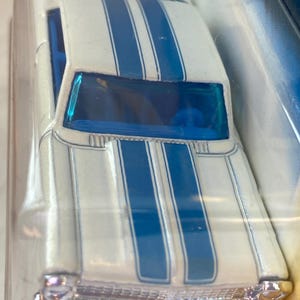 May include: A white die-cast toy car featuring two prominent blue racing stripes running along the hood and roof. The vehicle has a metallic silver front bumper, a bright blue windshield, and blue interior details visible through the clear plastic.