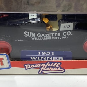 May include: A black toy race car with red wheels, featuring the text "SUN GAZETTE CO. WILLIAMSPORT, PA." and the number "137." The car is labeled "1951 WINNER" and is part of the "Downhill Heroes" collection.