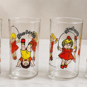 May include: Set of four clear drinking glasses featuring Cabbage Patch Kids illustrations. Each glass has a different image of a Cabbage Patch Kid playing with a jump rope. The glasses are clear with colorful graphics.