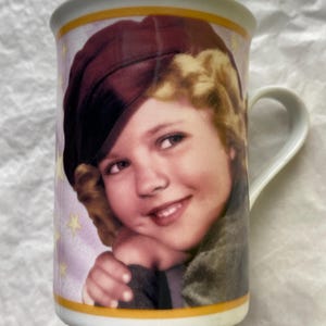 May include: A white ceramic mug featuring a printed portrait of a smiling child with blonde curly hair wearing a dark maroon beret. The background is a light purple color decorated with small yellow stars, framed by thin yellow bands at the top and bottom.
