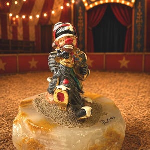 May include: A clown figurine in a circus setting. The clown is dressed in a black and gold outfit, with a red nose and a sad expression. The figurine is on a stone base. The background features a circus tent and stage.