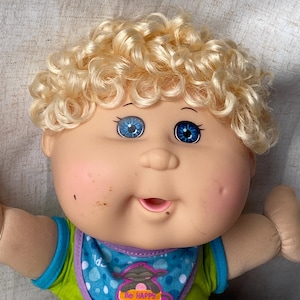 Op de afbeelding: Een pop met krullend blond haar, blauwe ogen en roze wangen. De pop draagt een groen shirt met blauwe randen en een blauwe slabbetje met een afbeelding van een aapje dat een bordje vasthoudt met de tekst Be HAPPY. De pop heeft een lichte huid.