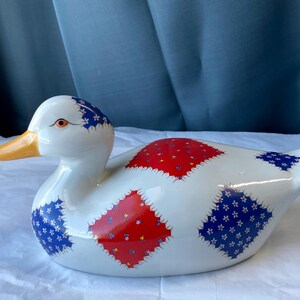 May include: A white ceramic duck figurine featuring a decorative pattern of red and blue diamond-shaped patches. Each patch is adorned with small floral motifs. The duck has a smooth, glossy finish, a bright orange beak, and a single visible eye.
