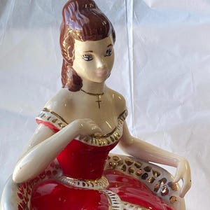 Puede incluir: Una figura de cerámica esmaltada de una persona con cabello castaño recogido, vistiendo un vestido rojo con ribetes dorados y un collar de cruz de oro. La figura está sentada, con detalles faciales pintados, ojos azules y un acabado brillante.