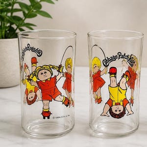 Può includere: Due bicchieri trasparenti con illustrazioni dei Cabbage Patch Kids. Uno mostra una ragazza con pantaloncini e top rossi che salta la corda, l'altro un ragazzo a testa in giù. Il testo "Cabbage Patch Kids" è stampato sui bicchieri.