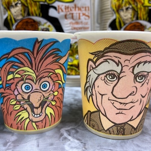 May include: Two disposable paper cups featuring colorful illustrations of fantasy characters. The left cup shows a shaggy, brown-furred creature with blue eyes on a blue background. The right cup displays a human-like character with a cap on a yellow background.
