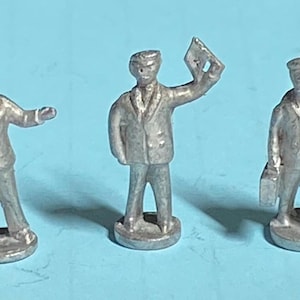 Puede incluir: Cinco pequeñas figuras de metal plateado de hombres en varias poses. Una figura está de pie con las manos juntas, mientras que otras gesticulan, sostienen un papel, llevan un maletín y señalan. Cada figura está sobre una pequeña base redonda.