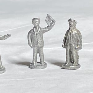 May include: Six small, silver-coloured metal figurines of people in various poses. One holds a briefcase, another gestures, and a third holds a paper. The figures appear to be wearing hats and coats, suggesting a vintage or historical theme.