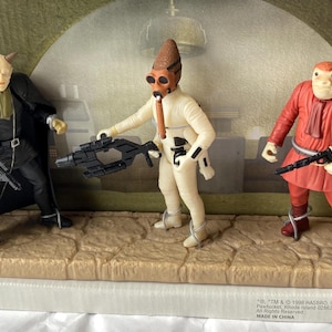 May include: A Star Wars action figure set featuring three characters. One figure is in a black outfit with a cape, another in a white outfit with a brown hat, and the third in a red outfit. Each figure is holding a weapon.
