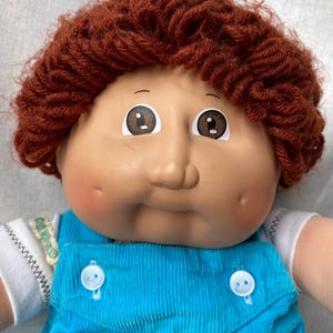May include: A doll with a round face, dimpled cheeks, and brown yarn hair. The doll features brown eyes and wears a white short-sleeved shirt under bright blue corduroy overalls with two white buttons on the front bib and a small green tag.
