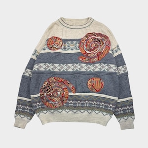 Op de afbeelding: Een vintage trui met een crèmekleurig bovenstuk en een blauw onderstuk. De trui heeft geometrische patronen en spiraalvormige patches in rood, oranje en bruin. De trui heeft lange mouwen en een ronde hals.