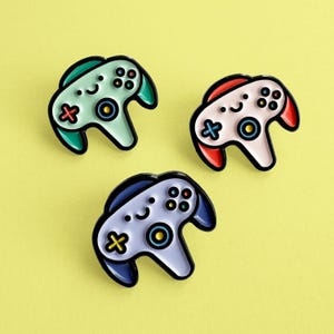 May include: Three enamel pins shaped like a video game controller. The controllers are in different colours: green, pink, and purple. Each controller has a smiley face and colourful buttons.