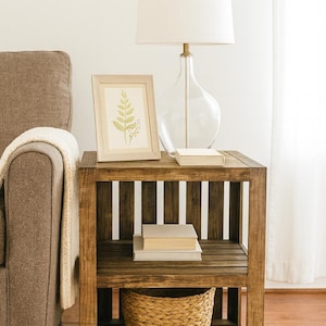 May include: A wooden side table with a framed botanical print, a clear glass lamp, and a woven basket. The table has two shelves with books on the top shelf. The lamp has a white lampshade. The table is next to a brown sofa.