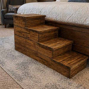 May include: A wooden pet step stool with four steps. The steps are stained a dark brown color and are constructed with visible wood grain. The step stool is placed next to a bed with a white comforter.