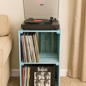 Puede incluir: Un tocadiscos negro con tapa transparente se encuentra sobre una estantería de caja azul claro. La estantería contiene discos de vinilo, incluido un álbum de los Beatles. La configuración está sobre una alfombra beige, con un sofá y una cortina beige en el fondo.