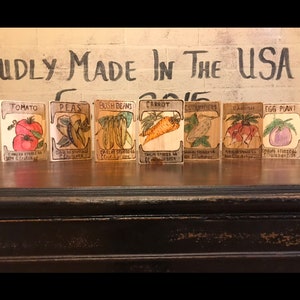 Puede incluir: Una colección de letreros de madera de verduras, cada uno con una ilustración pintada a mano y el nombre de la verdura. Los letreros incluyen tomate, guisantes, judías verdes, zanahoria, rábano y berenjena. Las palabras "PROUDLY MADE IN THE USA" están pintadas encima.