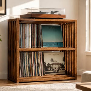 May include: A rectangular wooden record storage shelf with two open tiers featuring vertical slats on the sides. The unit is filled with vinyl records, including Fleetwood Mac's Rumours, and supports a dark wood turntable on the top surface.