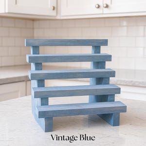 May include: A blue wooden display stand with five stepped levels. The stand is painted in a vintage blue color and has the words "Vintage Blue" printed at the bottom. The stand is designed to showcase items.