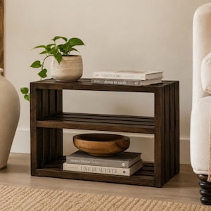 May include: A dark brown wooden two-tier shelf with a slatted design. The top shelf holds a potted green plant and two stacked books titled This is Home and down to earth. The bottom shelf holds a wooden bowl and two books titled monochrome home and LIVE BEAUTIFUL.