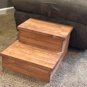 Puede incluir: Un taburete de madera de dos escalones con un acabado marrón rojizo cálido. El taburete está construido con tablones de madera, con cabezas de clavos visibles. El diseño es simple y funcional, adecuado para varios usos.