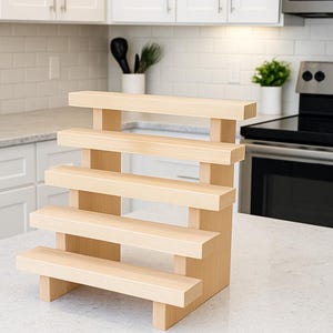 May include: A natural wood, multi-tiered display stand with five shelves. The stand is constructed from light-colored wood and features a simple, modern design. The text "Natural - No Stain" is visible at the bottom.