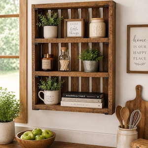 May include: A rectangular, dark brown wooden wall shelf with three horizontal tiers. The shelves hold small potted green plants, a white jar, a glass bottle with matches, a candle, and books titled Magnolia Table, Sunday Suppers, and The Kinfolk Table.