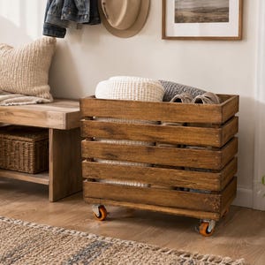 May include: A rectangular wooden storage crate with horizontal slats and a dark brown finish. The crate is mounted on four small orange swivel casters and contains folded white and gray textiles along with a pair of light-colored shoes.