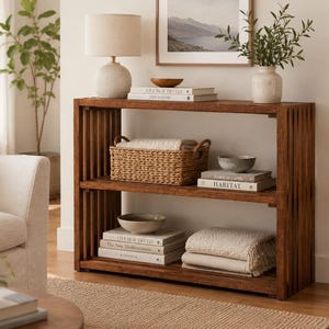 May include: A rectangular wooden console table with two open shelves featuring vertical slat side panels. The shelves hold a woven basket, stacks of books with visible titles, ceramic bowls, and folded cream-colored textile throws.