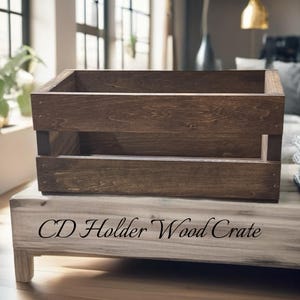 May include: A rectangular dark brown wooden crate with horizontal slats and an open top. The base features the text CD Holder Wood Crate in a black serif font. The crate is constructed from stained wood with visible grain and metal fasteners.