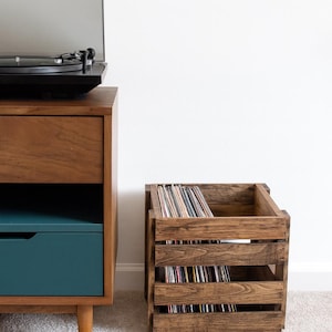 Peut inclure: Un coffre en bois rempli de disques vinyles est placé à côté d'un meuble de platine vinyle de style moderne du milieu du siècle. Le meuble a une finition en bois foncé avec un tiroir turquoise. Le coffre est en bois teinté.