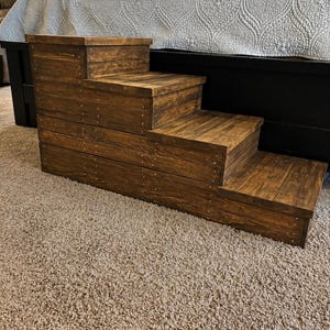 May include: A wooden pet step with four steps, stained a rich brown. The steps are constructed with visible wood grain and secured with small metal rivets. The pet step is designed to assist small dogs or cats in reaching a bed.