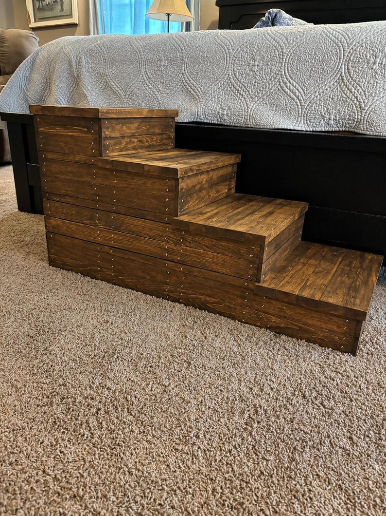 May include: A wooden pet step with four steps, stained a rich brown. The steps are constructed with visible wood grain and secured with small metal rivets. The pet step is designed to assist small dogs or cats in reaching a bed.