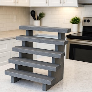 May include: A gray, tiered display stand with five levels. The stand is made of wood and has a textured finish. The text "Anique Gray" is visible on the bottom right. Ideal for displaying small items.