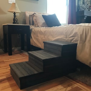 May include: Three-step pet stairs made of wood with a dark gray stain. The stairs are in front of a bed with a beige comforter and pillows. The stairs are on a hardwood floor.