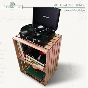 May include: A wooden record player stand with two shelves, holding a black record player and vinyl records. The stand is made of reclaimed wood and has a rustic finish. The record player is a Polaroid brand and has a black case with a silver handle. The stand is labeled "Studio 66" and "Rustic American Design, Handcrafted in Michigan".