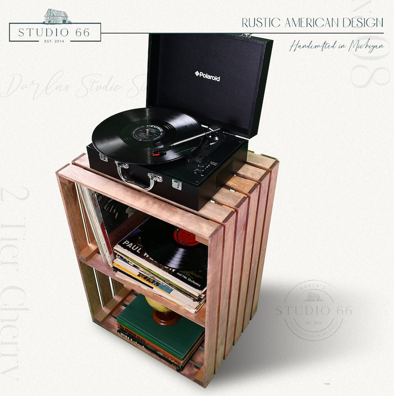 May include: A wooden record player stand with two shelves, holding a black record player and vinyl records. The stand is made of reclaimed wood and has a rustic finish. The record player is a Polaroid brand and has a black case with a silver handle. The stand is labeled "Studio 66" and "Rustic American Design, Handcrafted in Michigan".