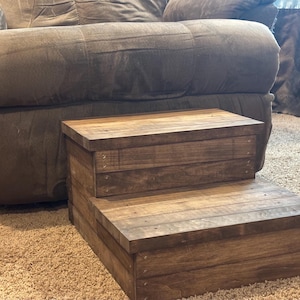 May include: A two-tier wooden pet step stool with a dark brown finish. The steps are rectangular and made of planks. The stool is on a light brown carpet in front of a grey sofa.