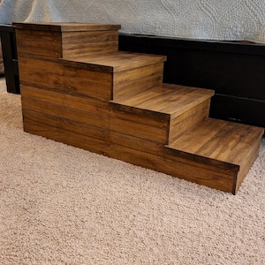 May include: A wooden pet step stool with three steps. The steps are made of stained wood and are positioned in front of a bed. The step stool is designed to help pets access higher surfaces.