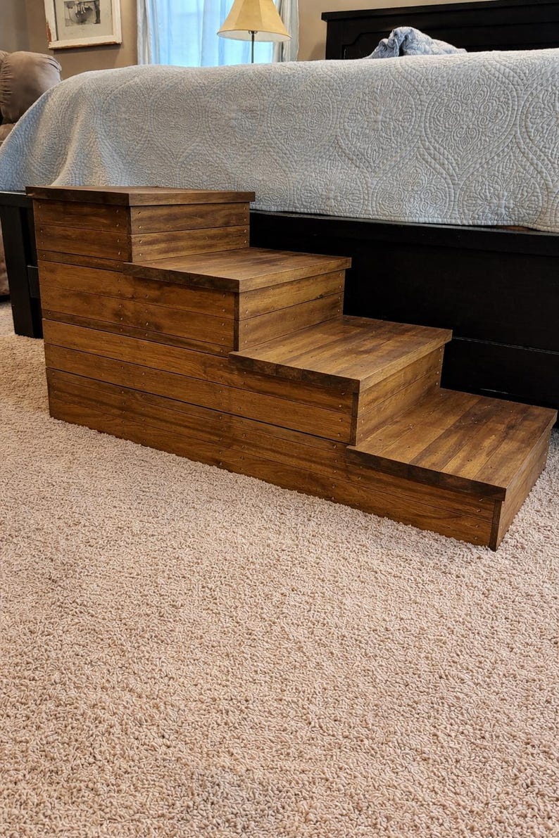 May include: A wooden pet step stool with three steps. The steps are made of stained wood and are positioned in front of a bed. The step stool is designed to help pets access higher surfaces.