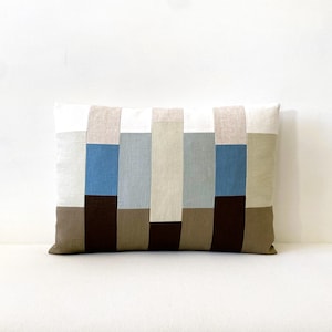 May include: Rectangular cushion with a patchwork design. It features vertical stripes in white, beige, blue, and brown. The cushion is made of soft fabric and is designed for home decor, measuring approximately 40cm x 60cm.
