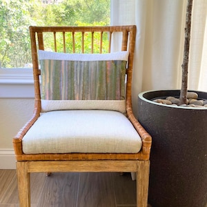 May include: A wicker chair with a rectangular pillow and a light-colored seat cushion. The pillow has a vertical striped pattern in shades of green, purple, and pink. The chair is next to a large dark pot with a tree.