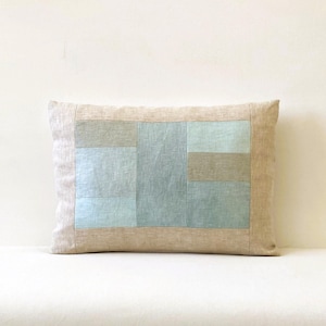 Peut inclure: Coussin rectangulaire décoratif avec un motif patchwork. Il présente une bordure beige clair et un panneau central composé de patchs rectangulaires dans des tons de bleu et de gris. Le coussin est posé sur une surface blanche.